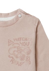 Lichtroze blouse met lange mouwen en een ronde halslijn met drukknoop. Voorzien van een grafisch ontwerp van een verrekijker en de tekst: 'IK KIJK JOU!'.