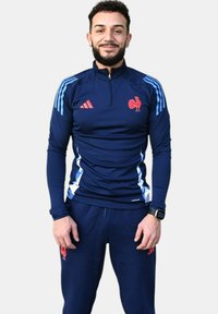 Chándal atlético azul que presenta una parte superior con media cremallera y acentos rojos, además de un logo de gallo. Incluye pantalones a juego con franjas laterales.