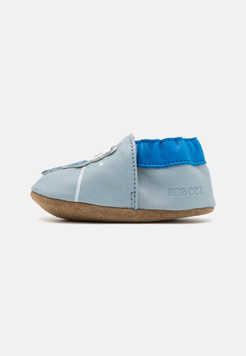 Zapatilla de cuero azul claro con parte superior elástica azul, suela de ante y detalles de costura; presenta un diseño decorativo en la parte frontal.