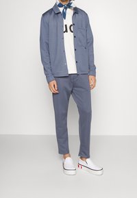 Conjunto casual de hombre que incluye una chaqueta azul claro, camiseta blanca con letras negras, pantalones azules a juego y zapatos blancos sin cordones con detalles en rojo.