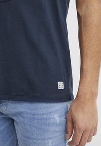 Marineblå bomulds T-shirt med et lille hvidt mærke, der står "DNM WORKRS" ved kanten. Stof er blødt med en glat tekstur.
