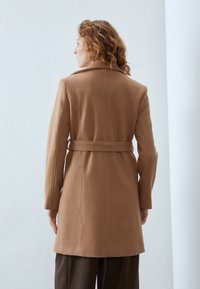 Beige ullblandingsfrakk med belte i midjen, lange ermer og spiss krage, med vertikale sømmer nedover ryggen for en strukturert design.