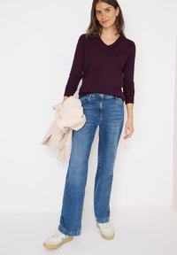 Paars V-hals sweater, blauwe flared jeans en crèmekleurige sneakers. Model houdt een beige fleecejack vast en staat tegen een witte achtergrond.