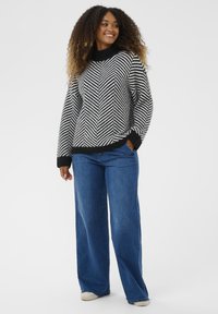 Femme souriante avec des cheveux bouclés, portant un pull en chevron noir et blanc, un jean large bleu et des baskets blanches, les mains dans les poches.