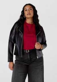 Zwarte leren jas met een asymmetrische rits, gedragen over een rood T-shirt. Donkergrijze hoge taillejeans en gouden accessoires maken de outfit compleet.