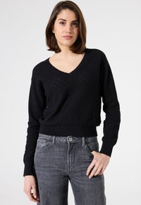 Pull noir en tricot avec un col en V et un motif texturé. Il possède des manches longues et une taille ajustée, associé à un jean gris.