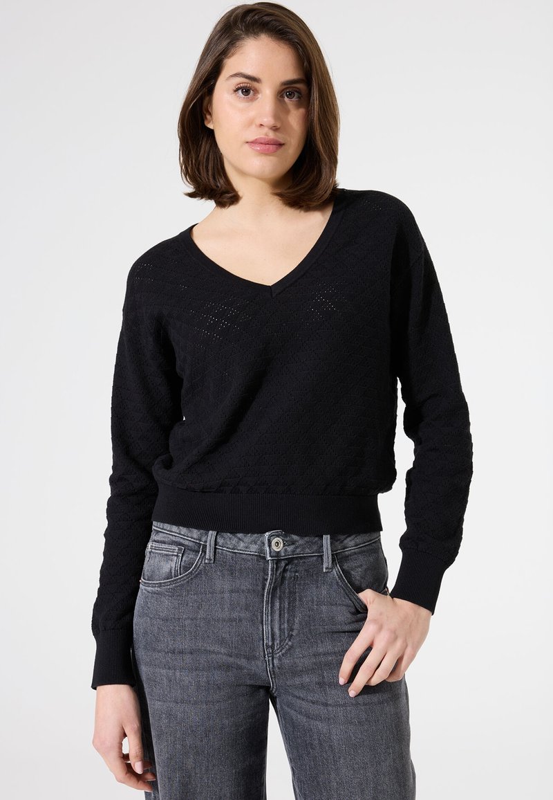 Pull noir en tricot avec un col en V et un motif texturé. Il possède des manches longues et une taille ajustée, associé à un jean gris.