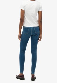 Vaqueros de mezclilla azul ajustados con una cintura de altura media, bolsillos traseros y un ligero estiramiento. Combinados con una camiseta blanca ajustada y zapatos oscuros.