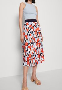 Top a manica larga di colore blu chiaro abbinato a una gonna midi plissettata con una stampa geometrica colorata in rosso, blu e bianco. Indossato con tacchi beige.