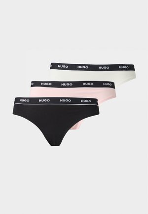 Jeune femme debout pieds nus portant un soutien-gorge vert olive et un slip noir avec une ceinture élastique sur un fond clair uni.