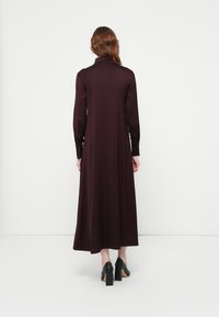 Robe bourgogne à manches longues avec col montant, texture lisse, design fluide et poignets boutonnés, associée à des chaussures à talons hauts noires.