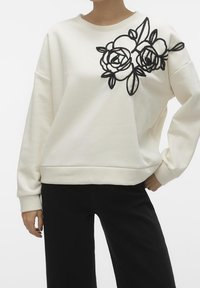 Cream-colored sweatshirt with long sleeves, featuring black embroidered roses on the left shoulder. Soft fabric with a relaxed fit.