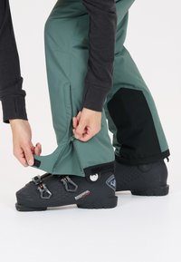 Gröna skidbyxor med dragkedjor vid fållen och svarta detaljer, i kombination med en svart skidstövel med justerbara spännen och strukturerad sula.