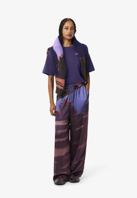 Top corto viola a maniche corte con logo, abbinato a pantaloni larghi a fantasia in viola e bordeaux. L'outfit è completato da uno scialle strutturato.