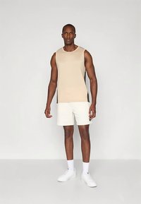 Top bege sem mangas com painéis laterais escuros em contraste, combinado com calções creme. Modelo em pé sobre um fundo neutro.