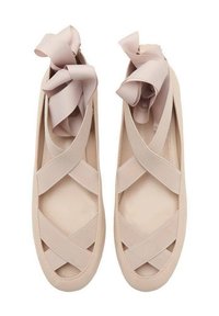 Chaussures plates beige avec des bandes de tissu croisées et de grands nœuds en ruban rose sur le dessus. Matériau doux et design à bout arrondi.