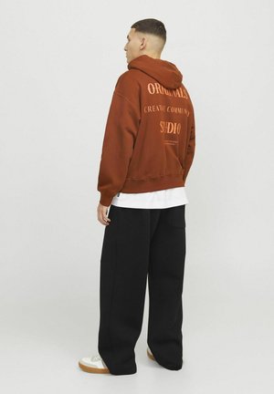 Brown hooded sweatshirt with orange text on the back, paired with black wide-leg pants. The sweatshirt features a relaxed fit and ribbed cuffs.
