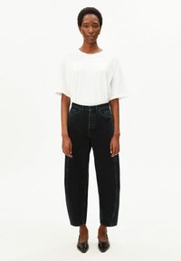 Femme debout face à l'avant, portant un t-shirt blanc uni oversize, un jean noir taille haute à jambes larges et des chaussures noires à bouts pointus.
