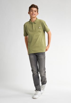 Olijfgroen polo shirt met een kraag, voorzien van een klein logo-patch. Gepaard met grijze jeans en witte sneakers, wat een casual uitstraling geeft.