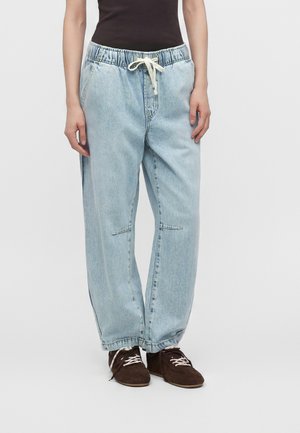 Persoană care poartă pantaloni de blugi largi, de culoare bleu deschis, cu talie elastică și șnur alb, asortați cu pantofi maro cu șiret.