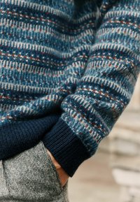 Blauer Wollpullover mit einem gemusterten Design aus Streifen und Punkten in Weiß und Rost, mit einem navyblauen gerippten Bund. Graue strukturierte Hose.
