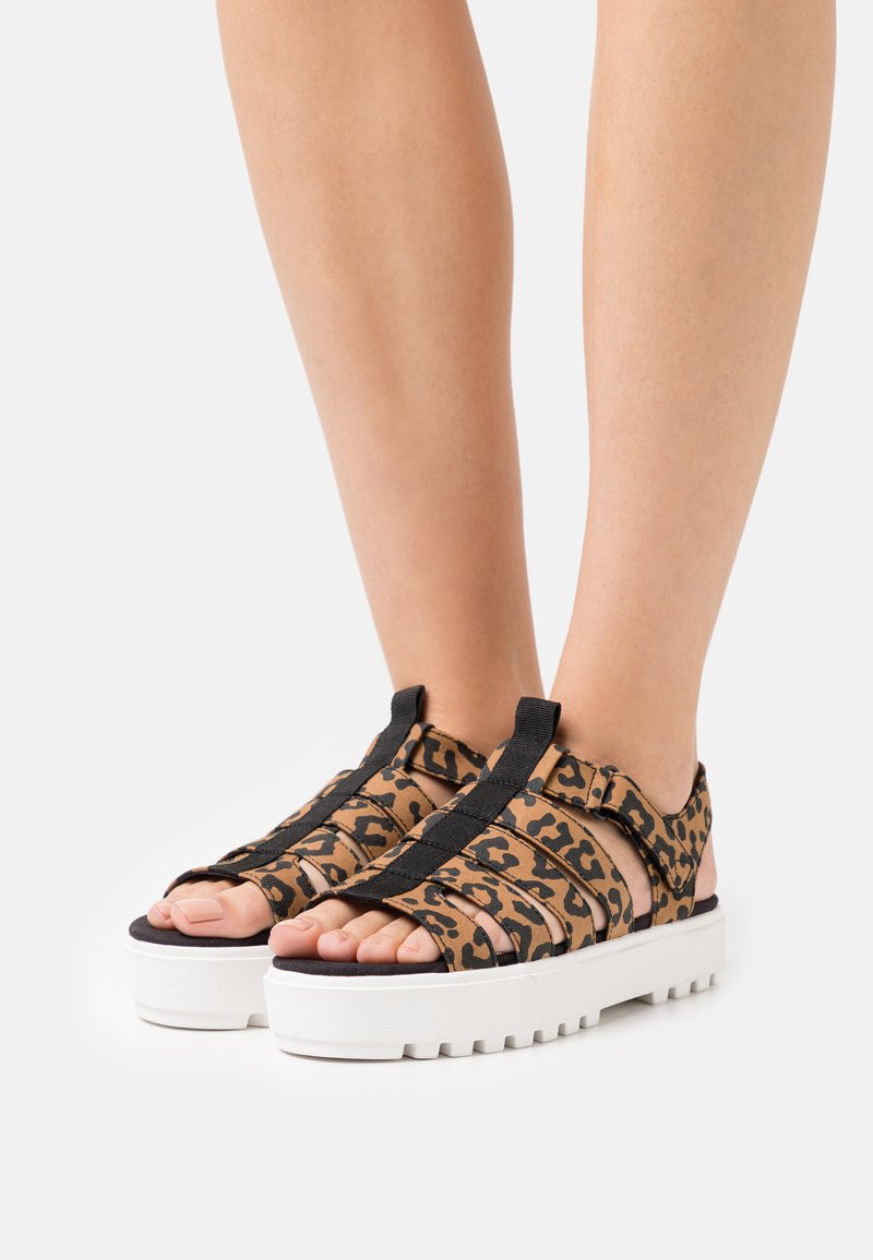 Sandálias com estampa de leopardo, com várias tiras pretas e uma sola branca espessa. Design aberto na frente que destaca os pés e os detalhes dos dedos.