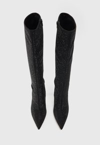 Bottes noires à talons aiguilles, arrivant jusqu'aux genoux, avec une surface texturée et scintillante. Elles présentent un bout pointu et une fermeture éclair à l'arrière.