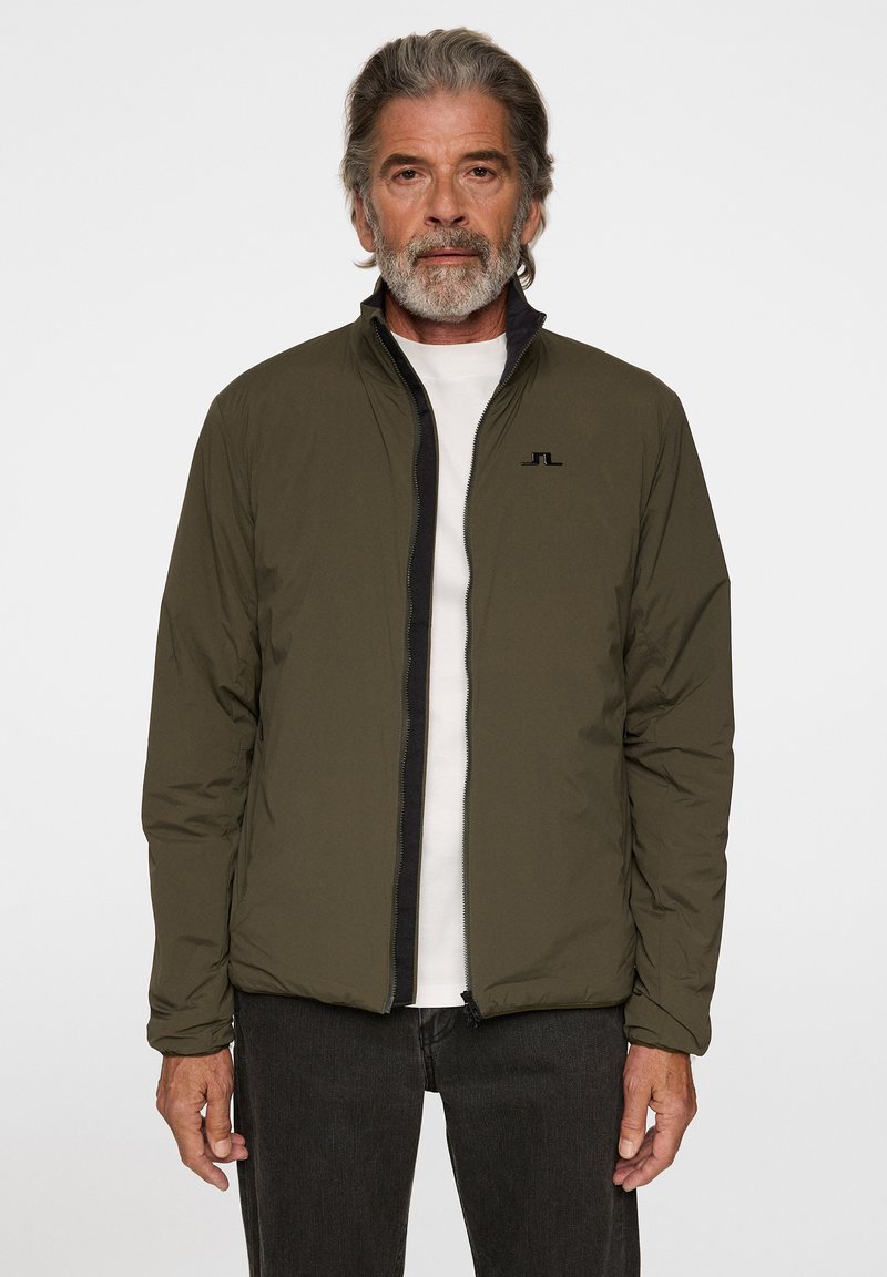 Olivgrön lättviktsjacka med svart dragkedja, hög krage och subtil logo på bröstet. Slen yta och minimalistisk design.