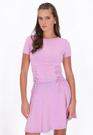 Tânără cu păr lung și castaniu, purtând un top liliac cu mâneci scurte și o fustă asortată cu detalii laterale cu șireturi, stând în fața unui fundal alb.