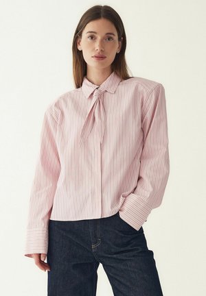 Vaaleanpunainen raidallinen paita, jossa solmittava kaulus, pitkät hihat ja napillinen etumukseen. Kankaassa pystysuoria valkoisia raitoja ja tekstuuri. Yhdistetty tummiin farkkuihin.