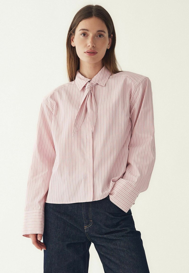 Růžová pruhovaná halenka s uvázaným límečkem, dlouhými rukávy a zapínáním na knoflíky. Texturovaná látka s vertikálními bílými pruhy. Kombinováno s tmavými džínami.