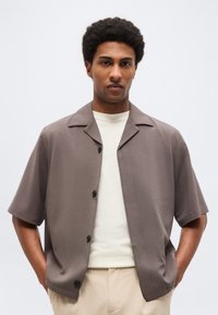 Άνδρας με κοντά σγουρά μαλλιά, φοράει ταουπί κοντομάνικο πουκάμισο με κουμπιά πάνω από λευκό μπλουζάκι και μπεζ παντελόνι, με τα χέρια στις τσέπες.