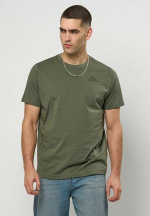 Muž má na sebe oličkovo zelené tričko s krátkym rukávom a malým logom na hrudi a svetlomodré rifle, pozerá sa doľava.