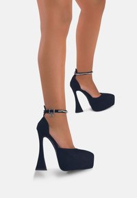 Scarpe con tacco platform nere in suede con punta arrotondata, alto tacco a stiletto e cinturino alla caviglia con hardware in argento e dettagli decorativi.