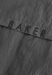 Szary materiał o lekko pogniecionej fakturze. Na górnej krawędzi widnieje odważny czarny napis "BAKER".