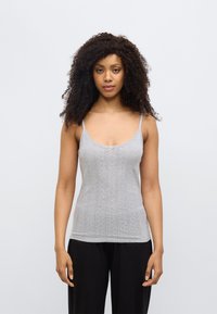 Débardeur gris en maille avec fines bretelles, présentant un motif texturé et un petit nœud au niveau de l'encolure. Porté avec un pantalon noir.