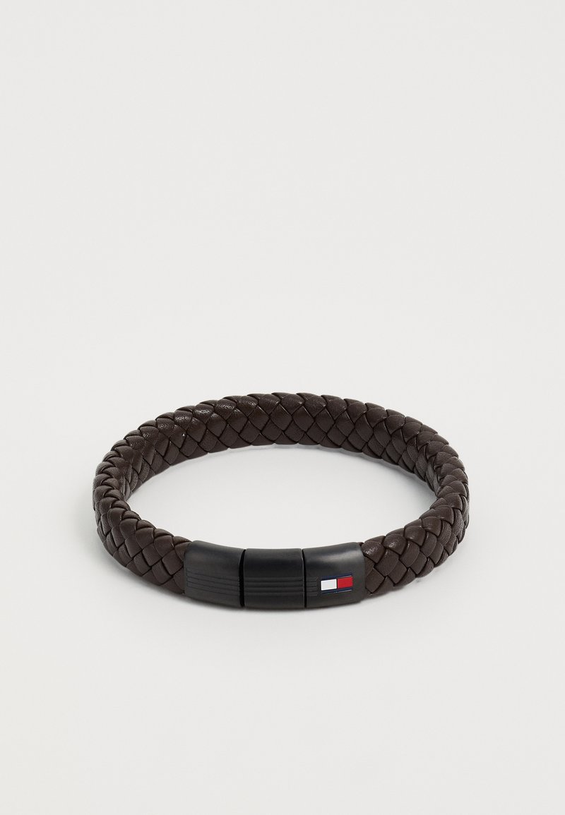 Pruun punutud nahast käevõru musta klambriga, millel on klambril väike logo punase, valge ja sinise värviga.