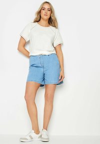 T-shirt grigio chiaro con cuciture decorative, abbinato a shorts di denim blu con vita elastica e tasche laterali. Sneakers bianche.