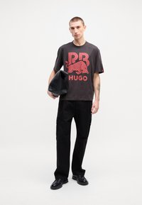 Black graphic t-shirt featuring red "PB HUGO" logo with a bull design. Paired with black cargo pants and holding a black shoulder bag.