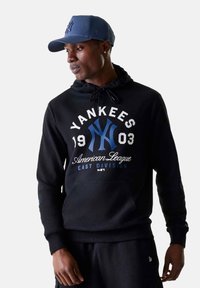 Felpa nera con tasca frontale, con la scritta "Yankees" e altro testo in bianco e blu. Abbinata a un cappellino da baseball blu. Tessuto liscio.