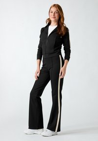 Femme portant une veste noire zippée et un pantalon assorti avec des bandes blanches sur les côtés, debout devant un fond clair uni.