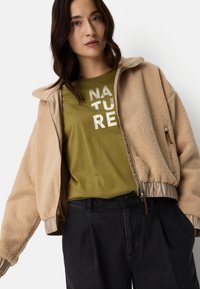 Beige fleece jacket with a light tan inner lining, featuring a zipper and elastic cuffs, worn over an olive-green shirt with metallic lettering.