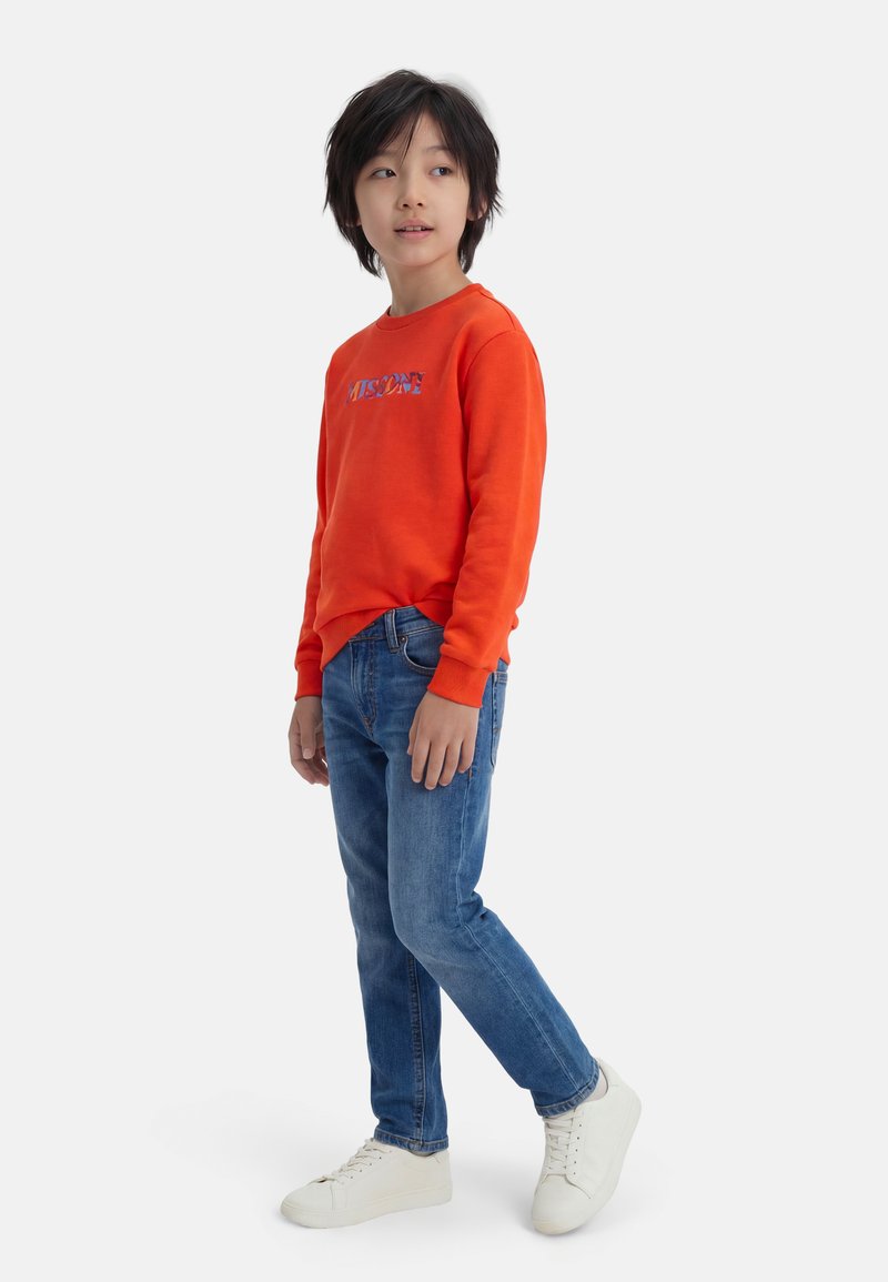 Enfant debout, portant un sweat-shirt orange avec un texte, un jean bleu et des baskets blanches, regardant légèrement sur le côté sur un fond uni.