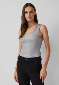 Vrouw met lang bruin haar, gekleed in een mouwloze zilveren tanktop en hoge zwarte broek, staande tegen een effen lichtgrijze achtergrond.