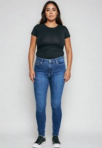 Žena stoji okrenuta prema naprijed, nosi crnu majicu kratkih rukava, plave uske traperice i crne tenisice s bijelim potplatom.