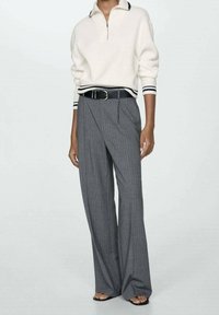 Modelo vistiendo una blusa blanca de manga larga con rayas negras en los puños y el cuello, pantalones anchos de cuadros grises, cinturón negro y sandalias negras.