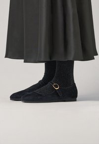 Bailarinas de ante negro con puntera redonda, correa con hebilla y una textura suave, combinadas con calcetines negros brillantes de canalé y una falda negra.