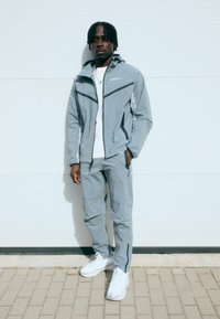 Gaiši pelēks sporta jaciņa ar melniem akcentiem, saskaņotas bikses un baltas kedas. Jaciņai ir rāvējslēdzējs un kapuce.