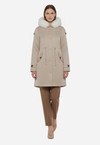 Parka beige con cappuccio in finta pelliccia bianca, vita con cordino, tasche laterali e bottoni a pressione. Abbinata a pantaloni beige e scarpe chiare.