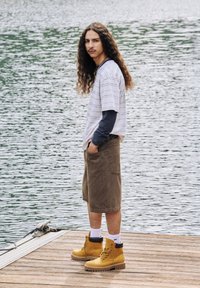 Brune snørebåndsstøvler, brune kordfløjlsshorts, hvid og marineblå stribet t-shirt, marineblå langærmet skjorte under, krøllet langt hår.